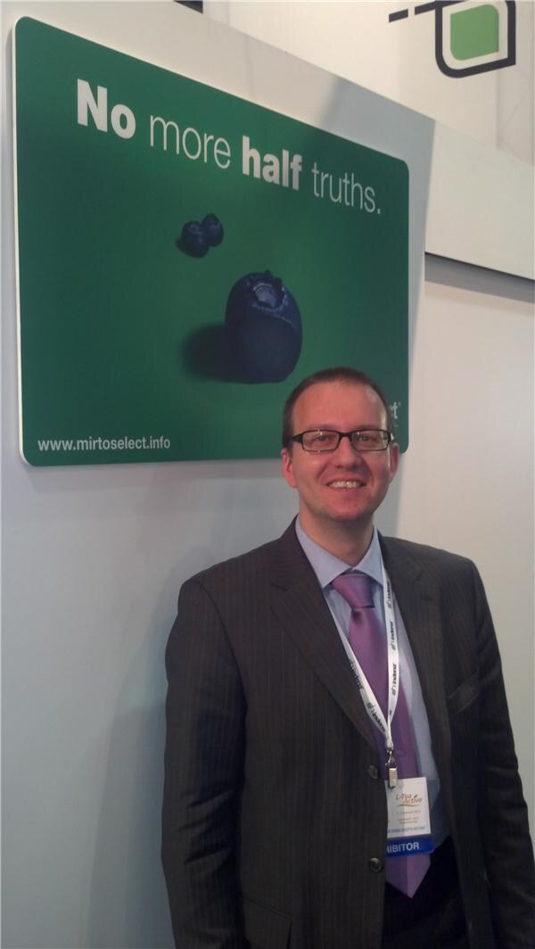 A smiling man with glasses, in a pinstriped suit and purple tie, stands beside a green Mirtoselect sign reading "No more half truths" with a blueberry image.