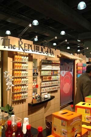 The Republic of Tea booth with wooden shelving of tea tins, a hanging banner and overhead lights; a person stands at the right and boxes sit in the foreground.