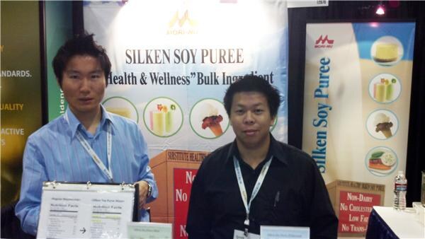 Two men wearing lanyards stand at a booth with Mori-Nu banners advertising Silken Soy Puree as a health and wellness bulk ingredient.