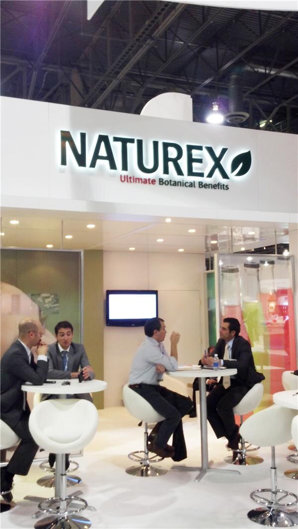 Naturex booth with an illuminated sign reading Ultimate Botanical Benefits. Four men in business attire sit and talk at two white round tables on white stools.