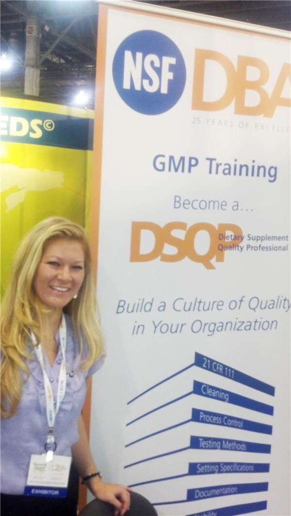 A smiling woman with long blonde hair and an exhibitor badge stands beside an NSF banner promoting GMP Training and becoming a Dietary Supplement Quality Professional.