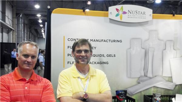 Two men with arms crossed stand in front of the NuStar Nutritional Manufacturing booth wall, which lists contract manufacturing and flexible packaging beside pouch shapes.