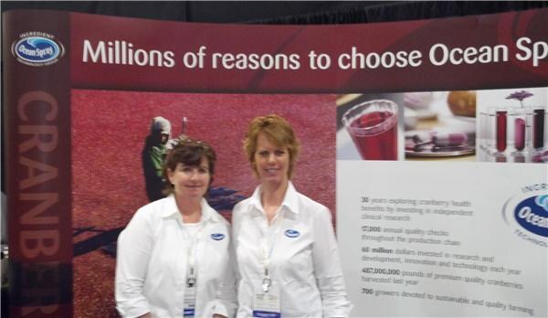 Two women in white Ocean Spray shirts stand in front of a cranberry-themed display reading Millions of reasons to choose Ocean Spray, with a glass of red juice pictured.