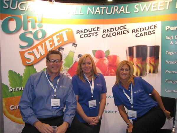 A man with glasses and two women in blue shirts and exhibitor badges pose in front of an Oh! So Sweet backdrop reading reduce costs, reduce calories, reduce carbs.