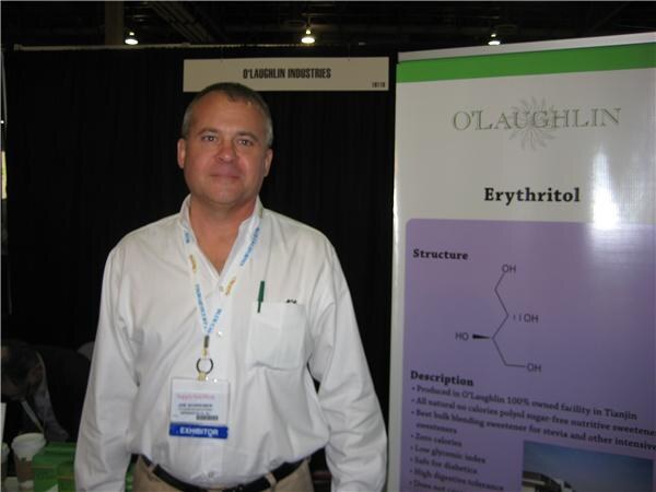 A man in a white shirt with an exhibitor badge stands in front of an O'Laughlin Industries sign and a banner about erythritol showing its chemical structure.