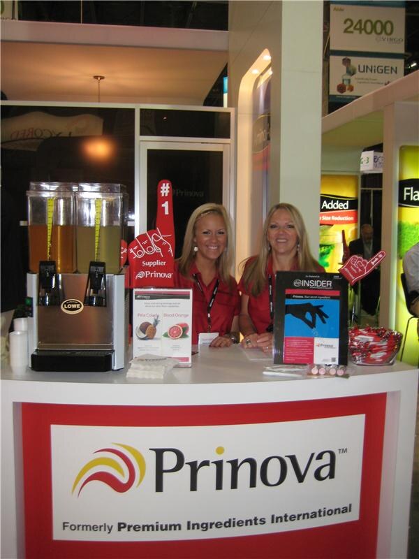 Two women in red shirts with lanyards stand behind a Prinova counter holding foam fingers. A Lowe dispenser with two orange drinks and a display card are on the counter.