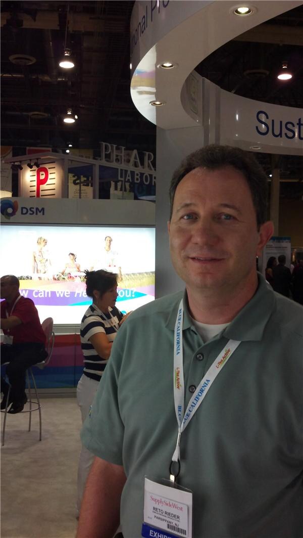 Reto Rieder, wearing a green polo and a SupplySide West badge, stands in the foreground in front of a DSM display with other people behind him.