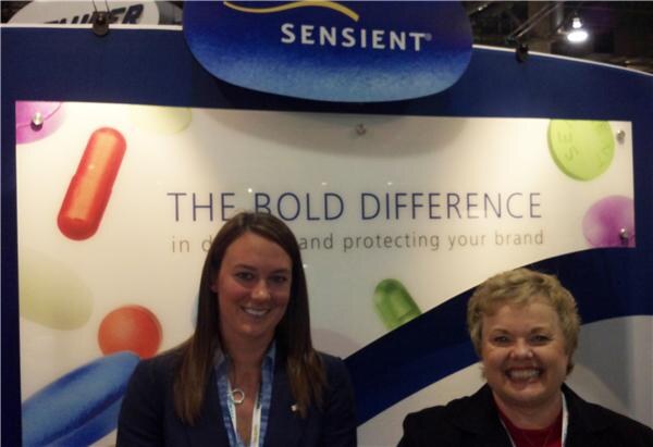 Two smiling women in dark jackets stand in front of a Sensient display reading The Bold Difference, decorated with colorful pill and capsule images.