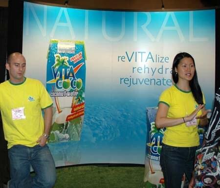 A man and a woman in yellow shirts stand in front of a Vita Coco coconut water backdrop reading Natural, revitalize, rehydrate, rejuvenate.