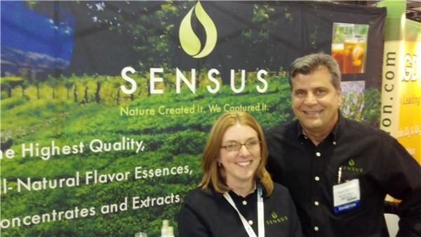 A smiling woman with glasses and a smiling man in black Sensus shirts stand in front of a green Sensus banner reading Nature Created It. We Captured It.