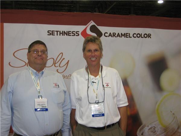 Two men with exhibitor badges stand in front of a Sethness Caramel Color banner showing a glass of cola with lemon slices; one wears a light blue shirt, the other a white polo.