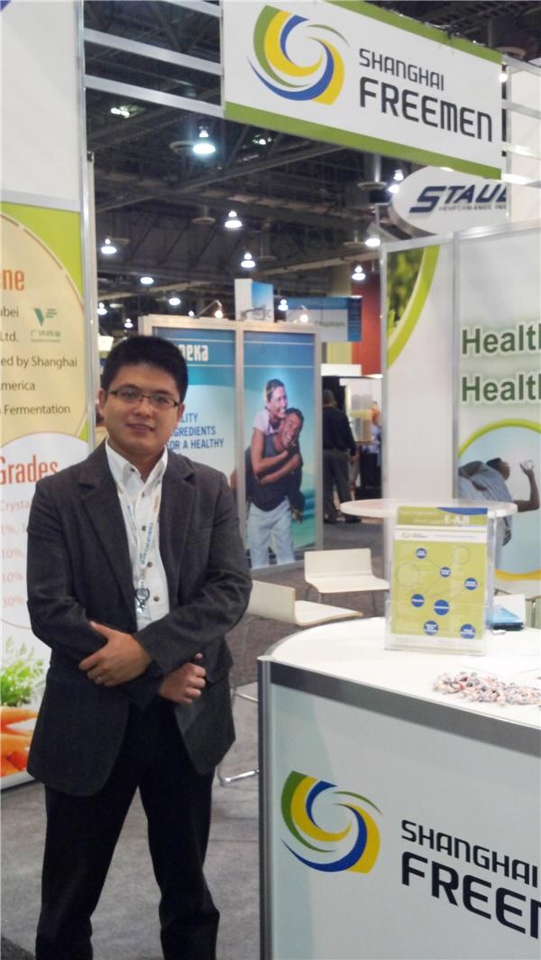 A man in a gray blazer and glasses stands beside a Shanghai Freemen display counter with brochures and candy, under a Shanghai Freemen sign.