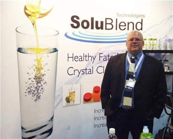 A man in a dark suit and striped tie with a lanyard stands in front of a SoluBlend Technologies banner showing oil being poured into a glass of clear water.