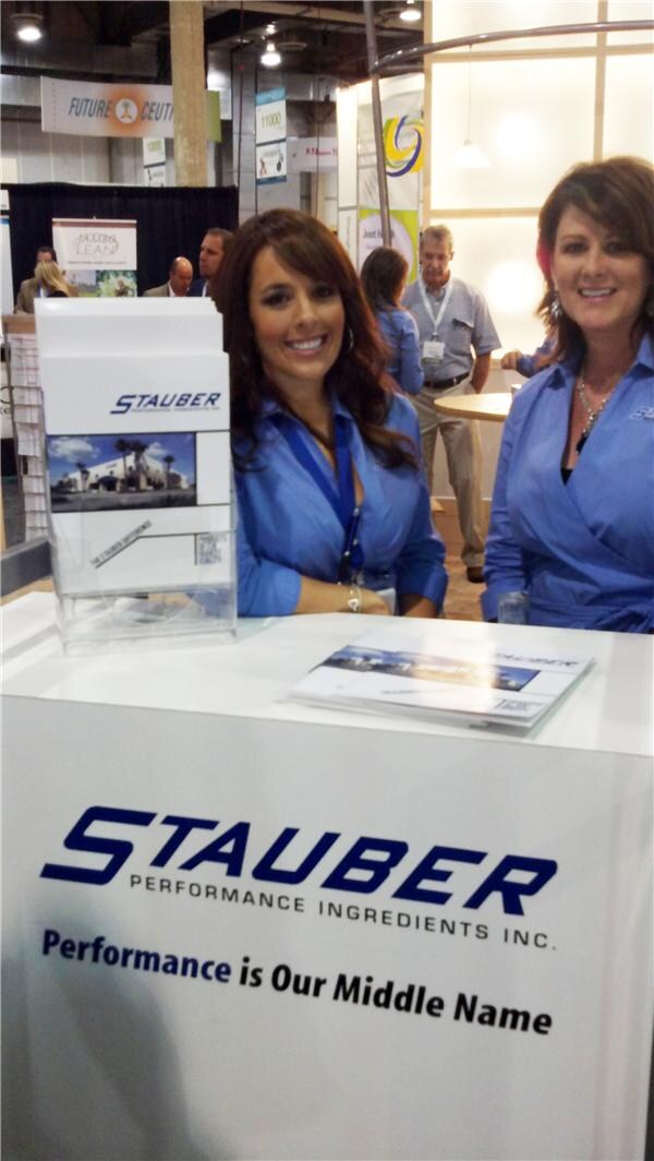 Two smiling women in blue shirts sit behind a white Stauber Performance Ingredients counter with brochures and a literature holder; other attendees are in the background.