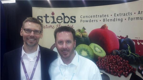 Two men stand close together in front of a stiebs banner showing pomegranate, kiwi and other fruit; one wears glasses and a dark jacket with a purple lanyard.