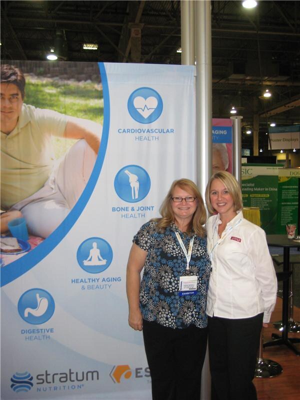 Two smiling women stand in front of a Stratum Nutrition banner listing health areas such as cardiovascular, bone and joint, and digestive health; one wears an exhibitor badge.