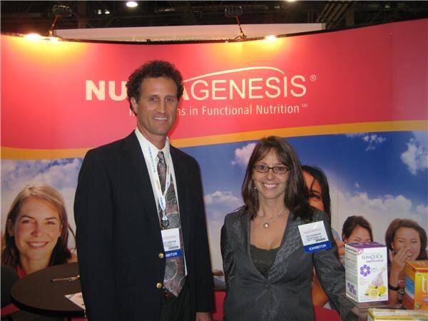 A man in a dark blazer and a woman with glasses, both wearing exhibitor badges, stand in a booth with a red banner. SubQuick product boxes sit on a table beside them.