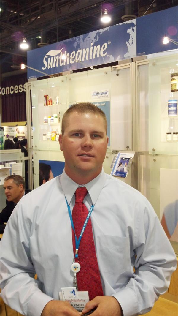A man in a light blue shirt and red tie with a blue lanyard stands in front of a Suntheanine booth with glass display cases holding product bottles.