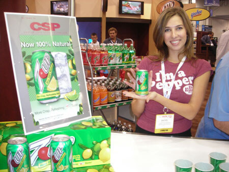 A smiling woman in a maroon shirt holds a can of 7UP beside a sign reading Now 100% Natural, with 7UP and A&W bottles on shelves and 7UP cartons in front.
