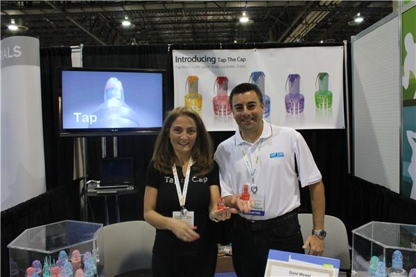A woman in black and a man in a white polo hold small colorful bottle caps in front of a screen and a Tap The Cap banner showing caps in several colors.