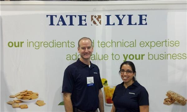 A man and a woman with glasses in navy polo shirts stand in front of a Tate & Lyle banner about ingredients and technical expertise, with crackers and a drink pictured.