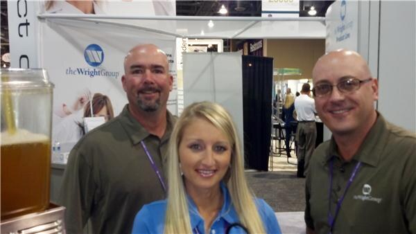 Two men and a blonde woman pose together at The Wright Group booth, with a company banner behind them and a drink dispenser at left. The men wear olive shirts and lanyards.