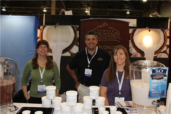 Two women and a man stand behind a sampling table with small cups at the Turtle Mountain booth. A So Delicious Vanilla Coconut Milk dispenser sits at right, and a Turtle Mountain banner hangs behind.