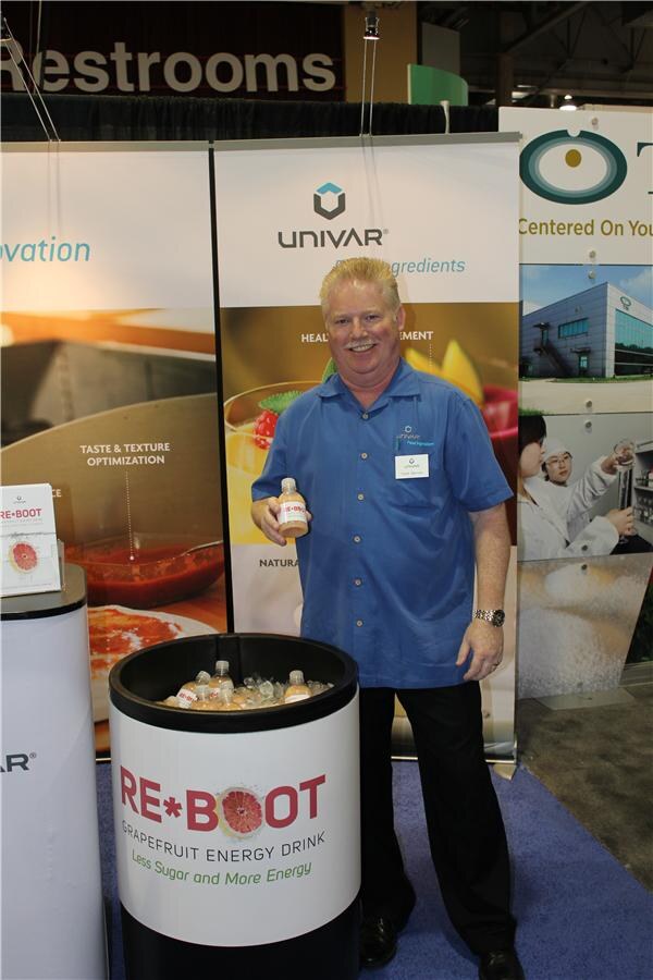 A smiling man in a blue shirt holds a bottle of RE*BOOT grapefruit energy drink beside an ice-filled bin of bottles, in front of Univar banners at the booth.