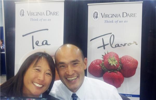 A smiling woman and man lean together in front of two Virginia Dare banners reading "Think of us as Tea" and "Think of us as Flavor," the latter showing strawberries.