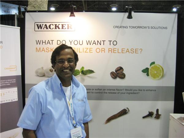 A smiling man with glasses and a light blue shirt stands in front of a Wacker display asking "What do you want to mask, stabilize or release?" with images of garlic, coffee beans and lemon.