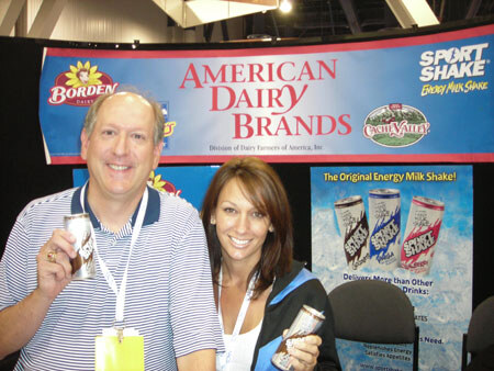 A smiling man in a striped polo and a smiling woman hold Sport Shake cans in front of an American Dairy Brands banner with Borden, Sport Shake and Cache Valley logos.