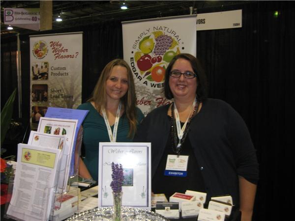 Two women stand behind a counter at the Weber Flavors booth, with brochures, a small vase of lavender and business cards in front of them and Weber Flavors banners behind.