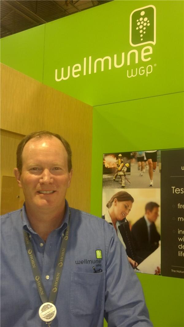 A smiling man in a blue Wellmune WGP shirt and lanyard stands in front of a green Wellmune WGP sign and a panel with photos of a firefighter, runners and office workers.