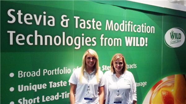 Two women in white shirts stand in front of a green WILD banner reading "Stevia & Taste Modification Technologies from WILD!"
