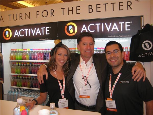 A woman and two men, arms around each other, smile at the Activate booth. Behind them are coolers of colorful Activate drink bottles under Activate signs.
