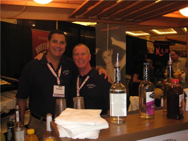 Two smiling men in black shirts and lanyards stand with arms around each other behind a bar counter holding bottles and napkins. A Kah banner is visible in the background.