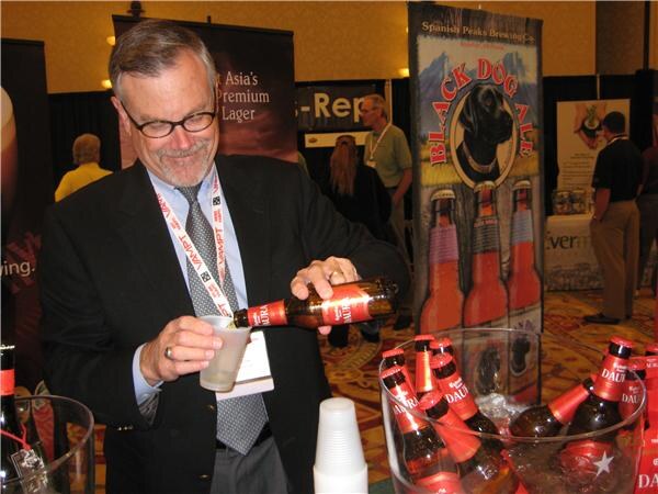 A smiling man with glasses and a lanyard pours a bottle of Estrella Damm beer into a plastic cup. Bottles sit in ice in the foreground, with a Spanish Peaks Brewing Co. Black Dog Ale banner behind him.