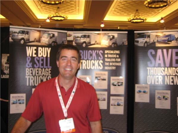A smiling man in a red polo and lanyard stands in front of Buck's Trucks display panels reading "We buy & sell beverage trucks" and "Save thousands over new," with truck photos.