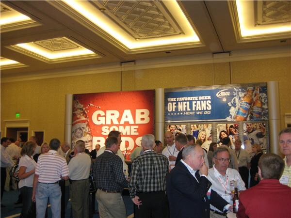 Attendees stand and talk in a large ballroom in front of Bud Light signs, one reading "Grab some Buds" and another "The favorite beer of NFL fans." One man holds a bottle.