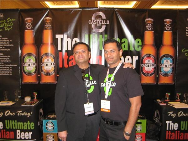 Two men in dark clothing and badges pose with arms around each other in front of a Castello lager beer banner showing four beer bottles and the words "The Ultimate Italian Beer."
