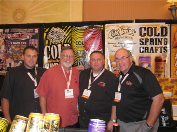 Four men with badges stand together at a booth with Cold Spring Brewing Company and Ol' Glory banners behind them and beverage cans on the counter in front.
