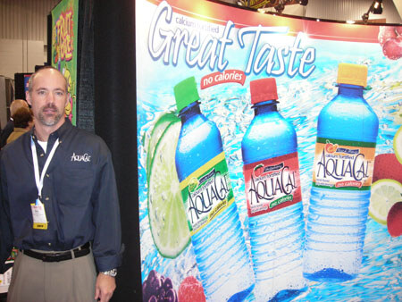 A bearded man in a dark AquaCal shirt and badge stands beside a large display reading Great Taste, no calories, showing three AquaCal calcium-fortified water bottles with fruit.
