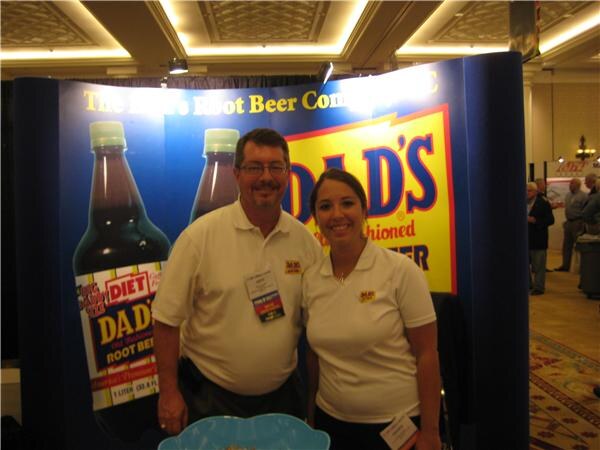 A man with glasses and a woman, both in white polo shirts with Dad's logos, stand in front of a Dad's Root Beer display showing large bottles and a yellow sign.