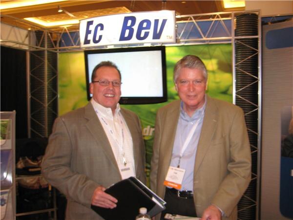 Two men in tan blazers and lanyards stand smiling in front of an EcoBev sign and a monitor in a truss-framed booth; one holds a black folder.