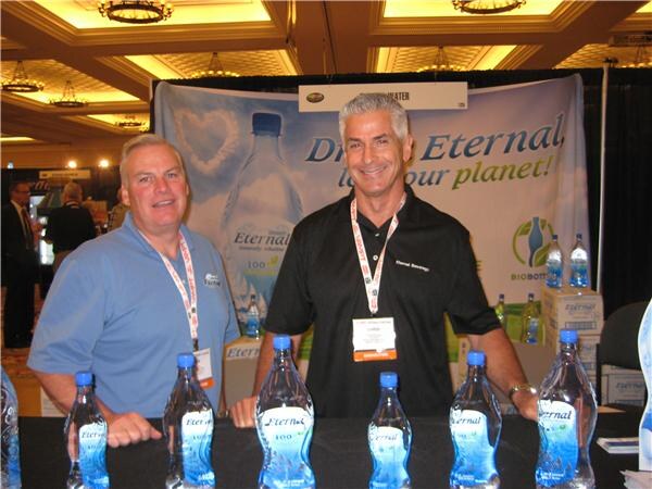 Two men with lanyards stand behind a counter lined with Eternal water bottles, in front of a banner reading "Drink Eternal" with a large bottle graphic.