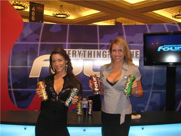 Two women hold cans of Four Loko in front of a purple camouflage-style backdrop with a large Four Loko logo. A television screen stands to the right, and more cans sit on the counter.