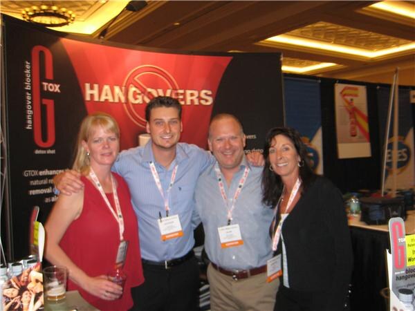 Four people with lanyards, two women and two men, pose with arms around each other in front of a GTOX hangover blocker banner reading "Hangovers" with a crossed-out symbol.