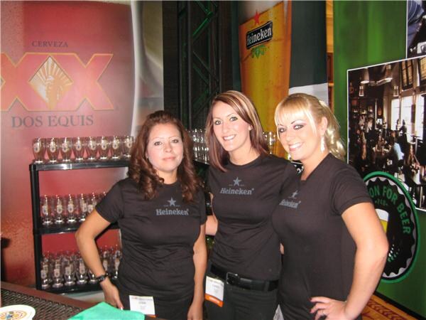 Three smiling women in black Heineken t-shirts stand at a bar with shelves of glasses, in front of a Dos Equis backdrop and Heineken banners.