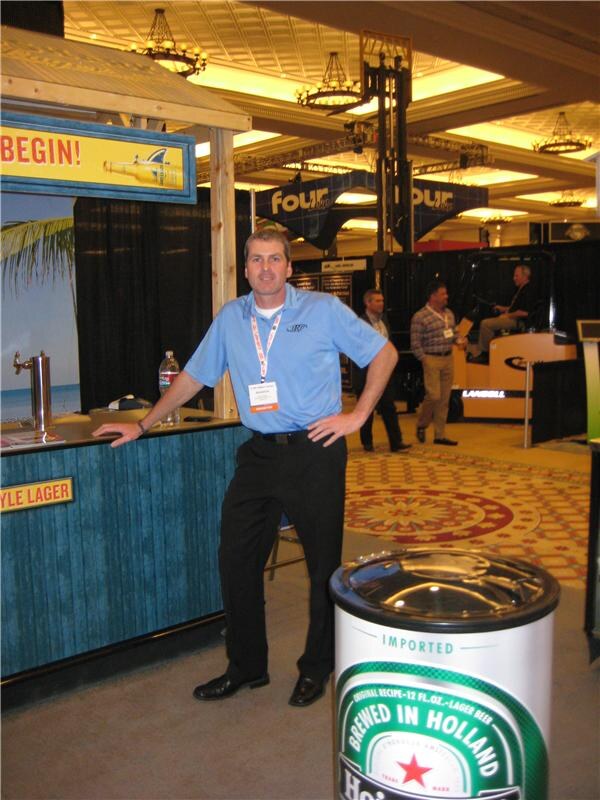 A man in a blue polo and badge leans on a tiki-style bar counter next to a Heineken-branded cooler marked Brewed in Holland, with other booths behind him.