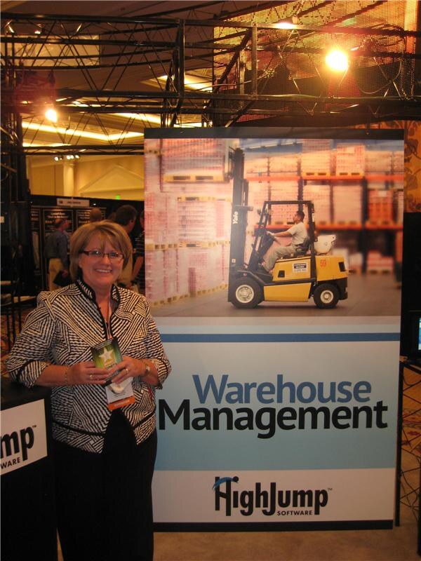 A smiling woman with glasses and a badge stands beside a HighJump Software Warehouse Management sign showing a yellow forklift in a warehouse.
